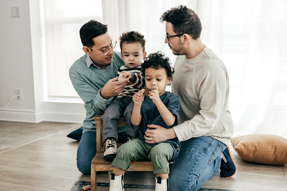 Two fathers and two young boys sitting on a chair engaged in conversation